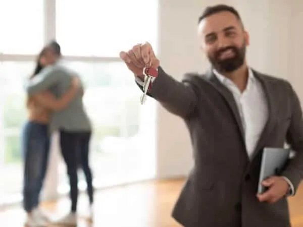 A real estate agent smiling and holding a key with a couple of tenants embracing each other in the background.