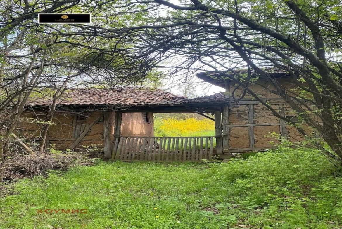 Village house from a plot for sale in the village of Ochusha, with green grass and trees around it. The house looks old with tiled roof and wooden fence.
