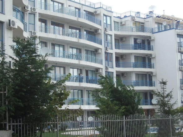 Модерен жилищен блок със светли фасади и сини балкони, заобиколен от зелени дървета и ограден с метална ограда.