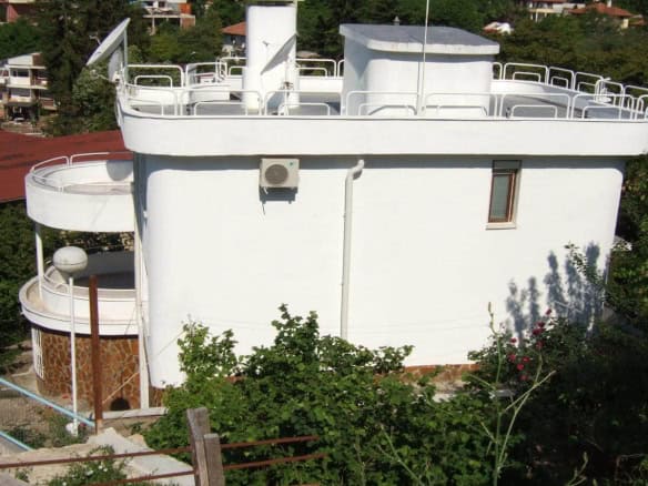 A peculiar white, round building with a flat roof, surrounded by white railings and equipped with several satellite dishes, set amidst green bushes.
