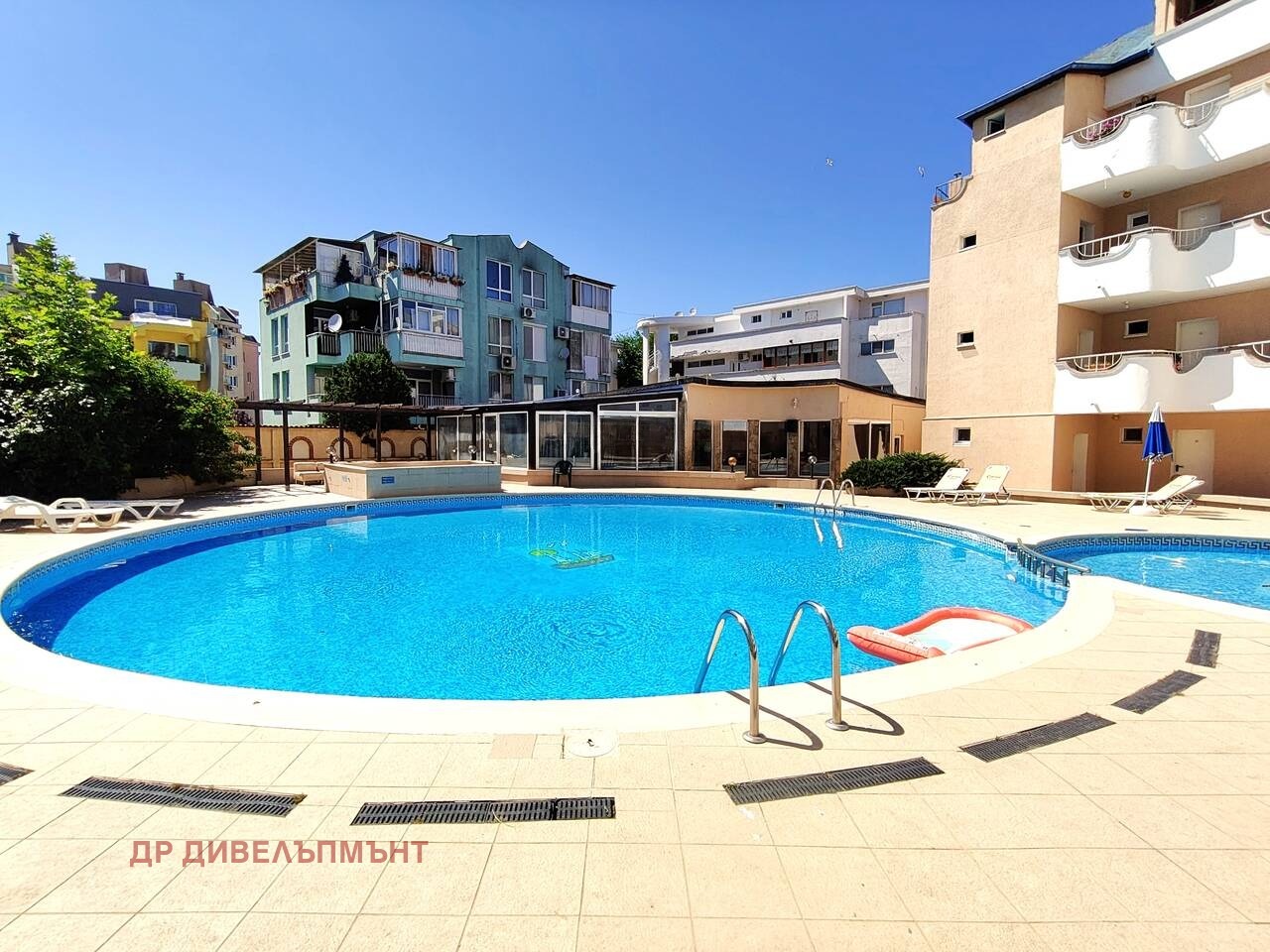 Large oval outdoor swimming pool with ladder, sun loungers, and adjacent building