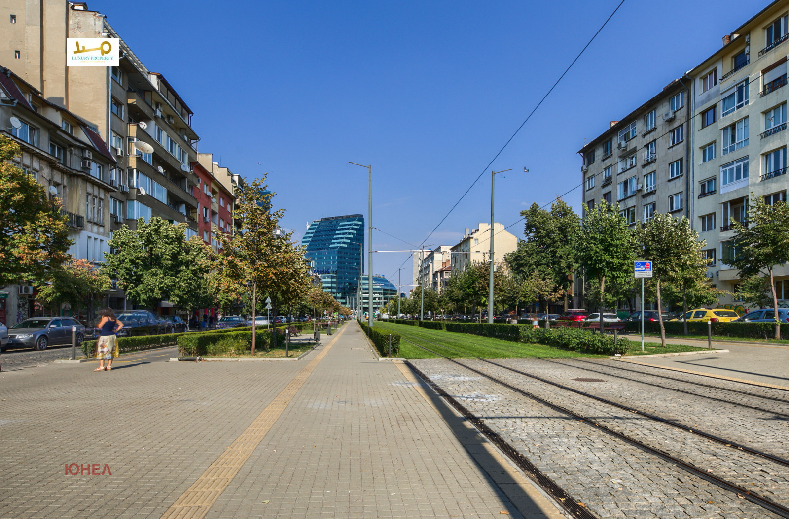 Wide boulevard with tram tracks, tree-lined median, pedestrian pavement, modern glass building in background
