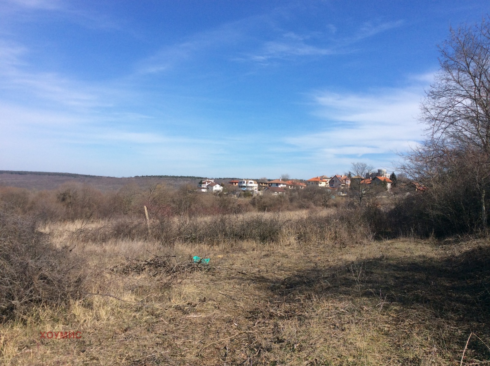 Open land plot with dry grass, bare shrubs, and village in background
