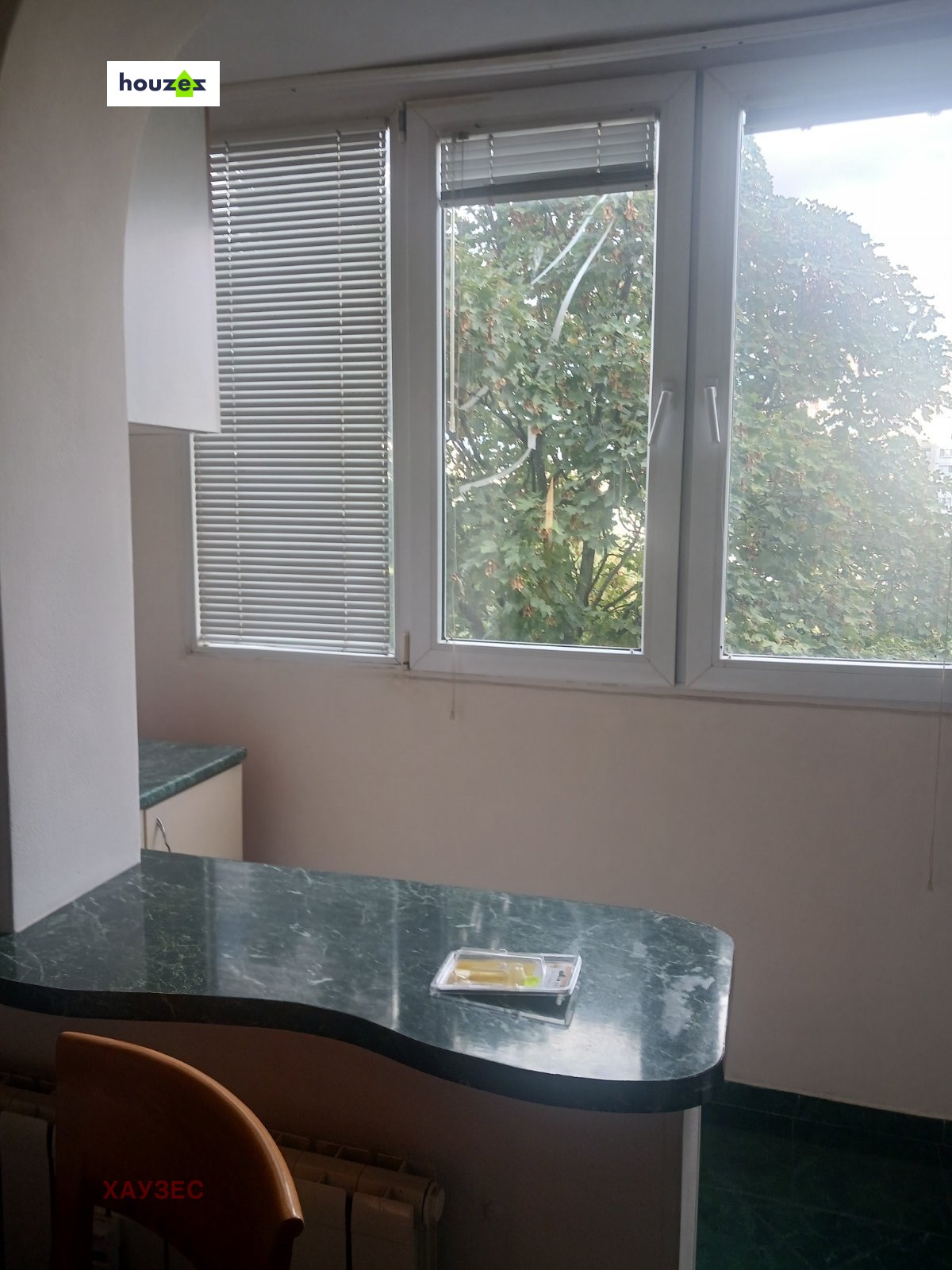 tristaen-apartament-90-sqm-pod-naem-lyulin-3-sofia-obzaveden-sklad-7 Кухненска трапезария със зелена мраморна маса, прозорец с щори и изглед към дървета