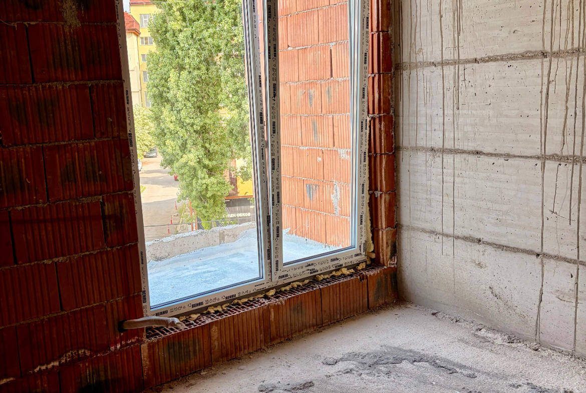 tristaen-apartament-108-sqm-za-prodazhba-ovcha-kupel-novo-stroitelstvo-6 Недовършена ъглова стая с наклонен таван, голям прозорец и изглед към дървета и улица