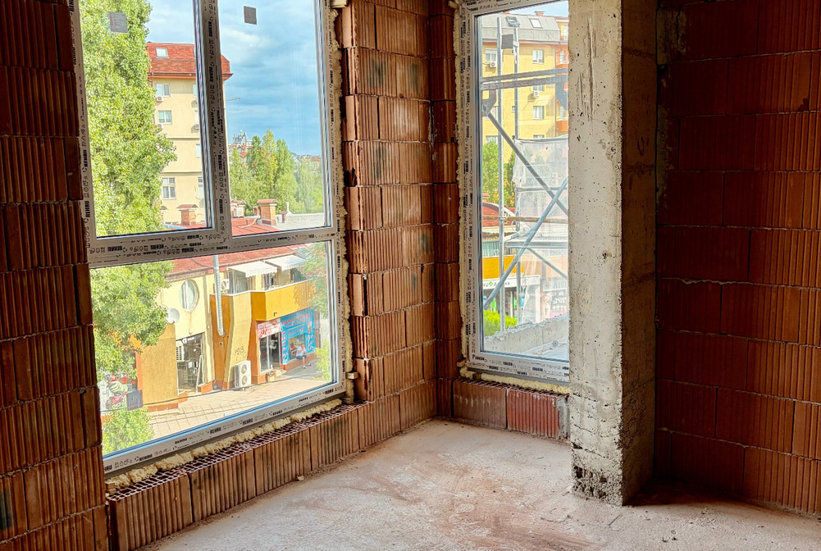 tristaen-apartament-108-sqm-za-prodazhba-ovcha-kupel-novo-stroitelstvo-8 Недовършена стая с два ъглови прозореца с изглед към улица, дървета и съседни сгради