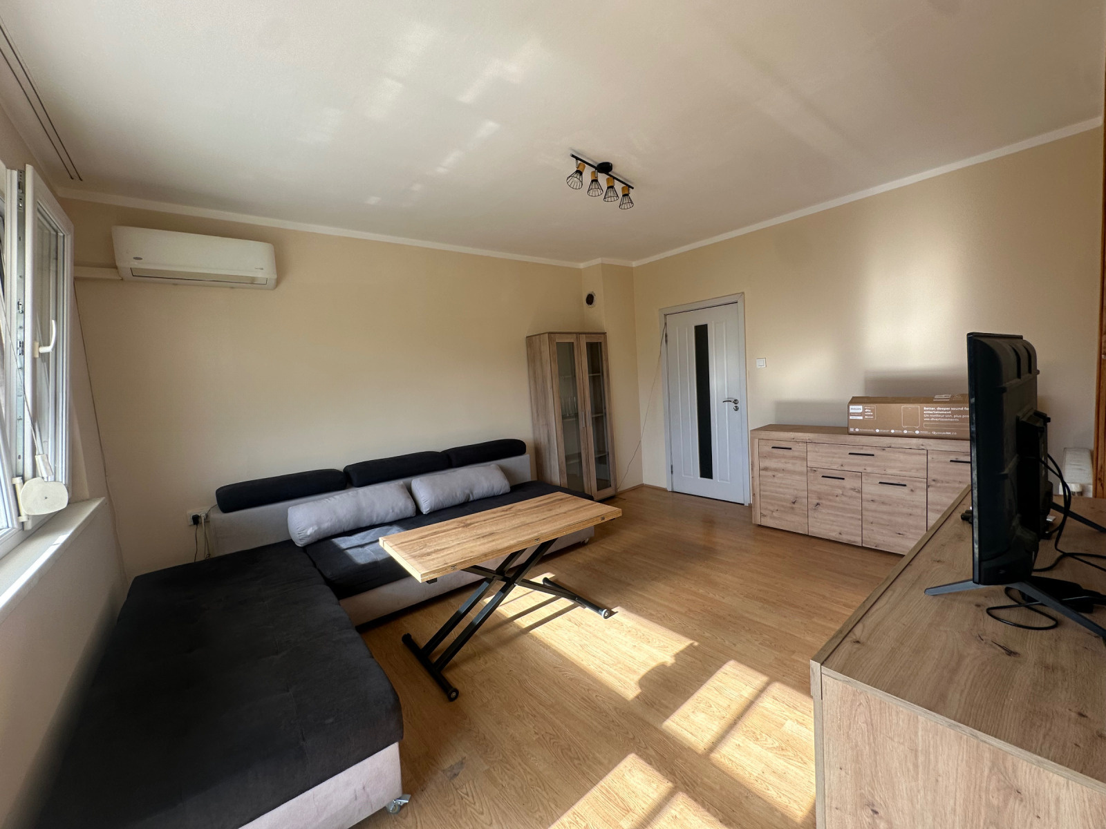 Дневна стая, гледана от срещуположния ъгъл с диван, повдигаща се маса, скрин, телевизор и бяла врата
